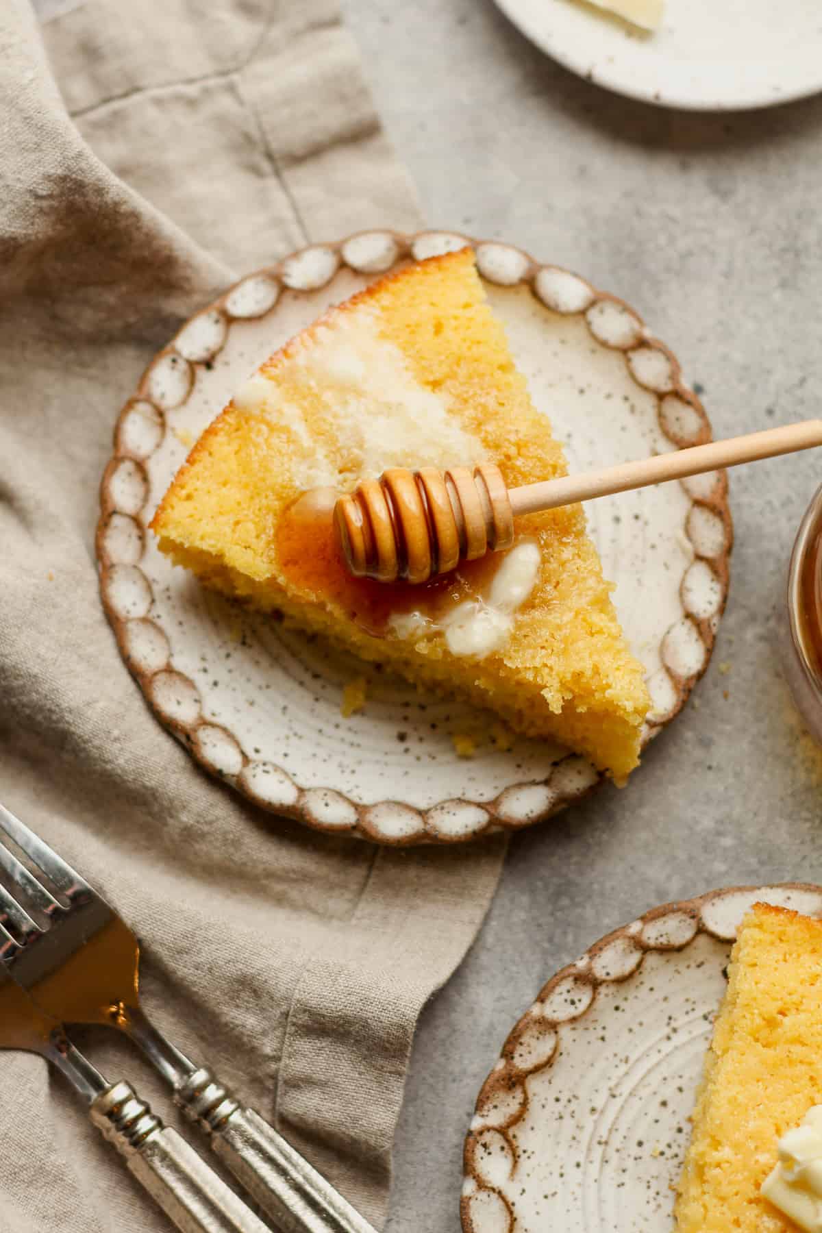 A small plate of a wedge of sweet cornbread with butter and a honey tool.