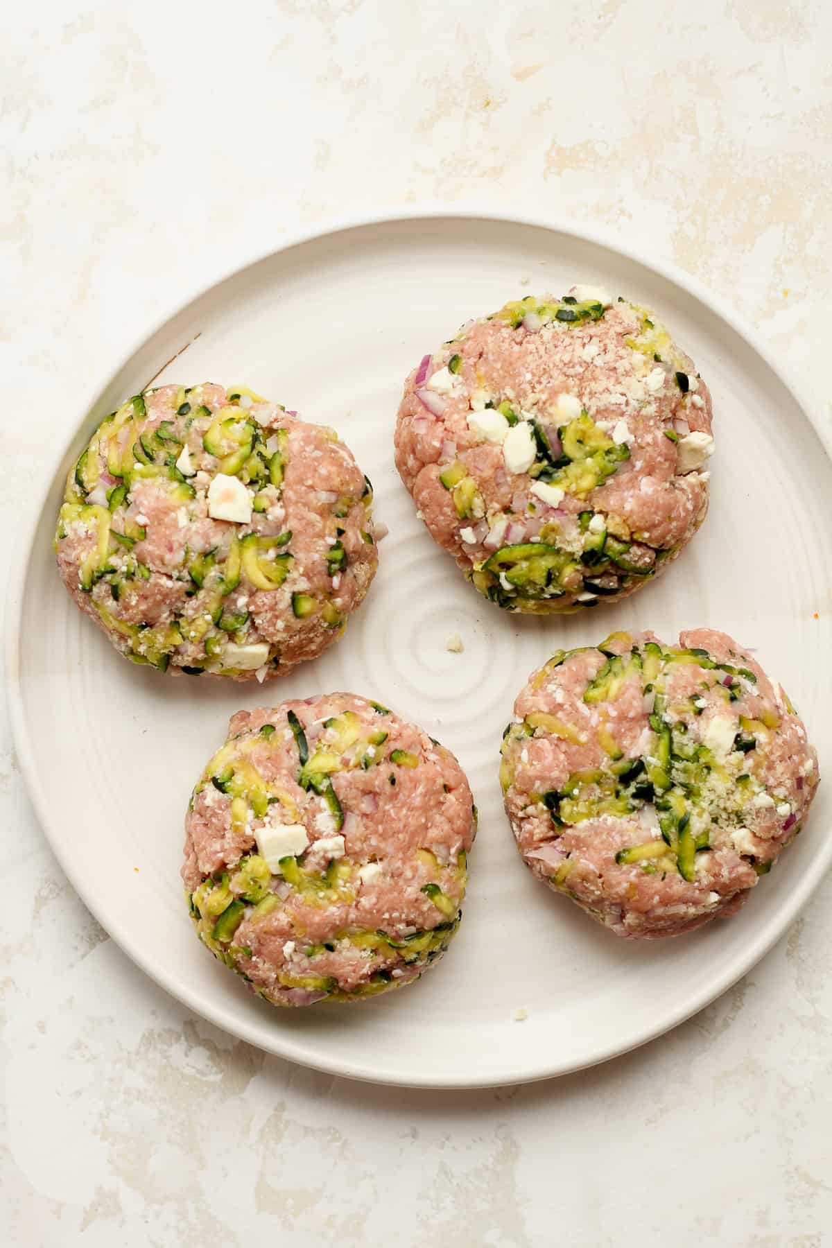 Overhead view of a plate of raw turkey burgers with zucchini and feta cheese.