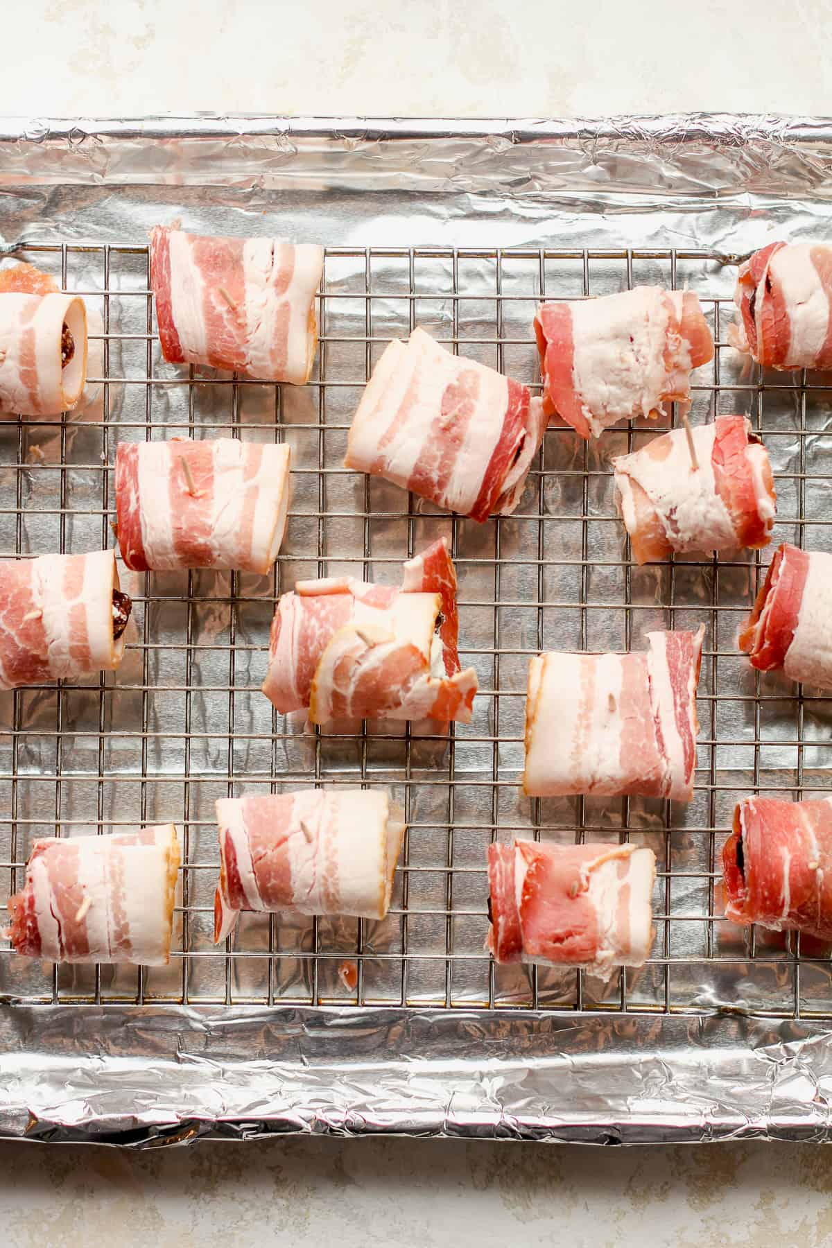 Some dates stuffed with goat cheese wrapped in bacon on a wire rack on top of a baking sheet.