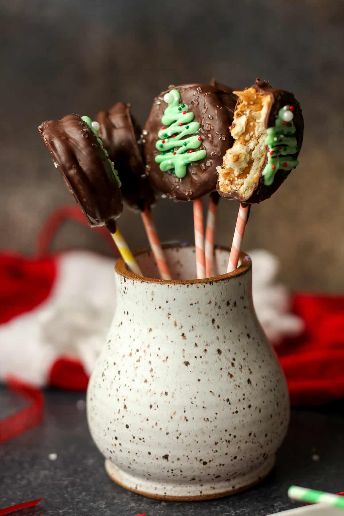 A mug with some chocolate covered ritz pops with Christmas trees drawn on.
