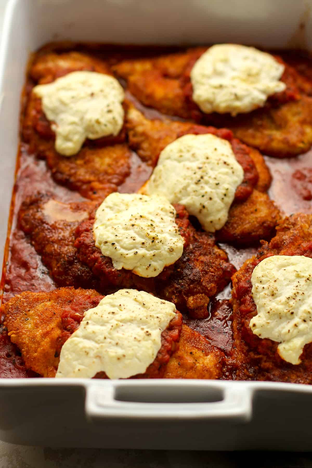 A casserole of six pieces of chicken parm with ricotta cheese on top.