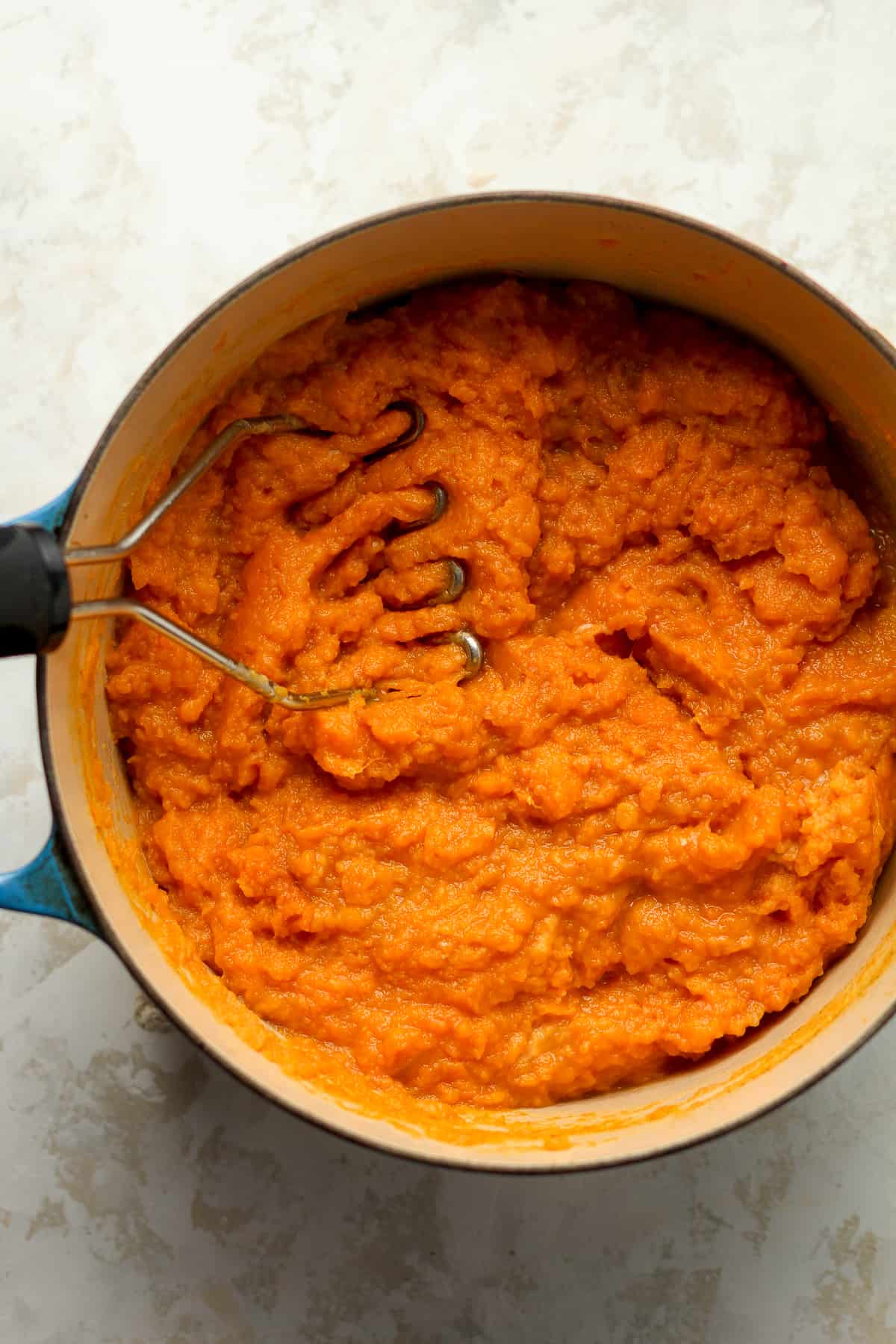 A stock pot of mashed sweet potatoes with a masher inside.