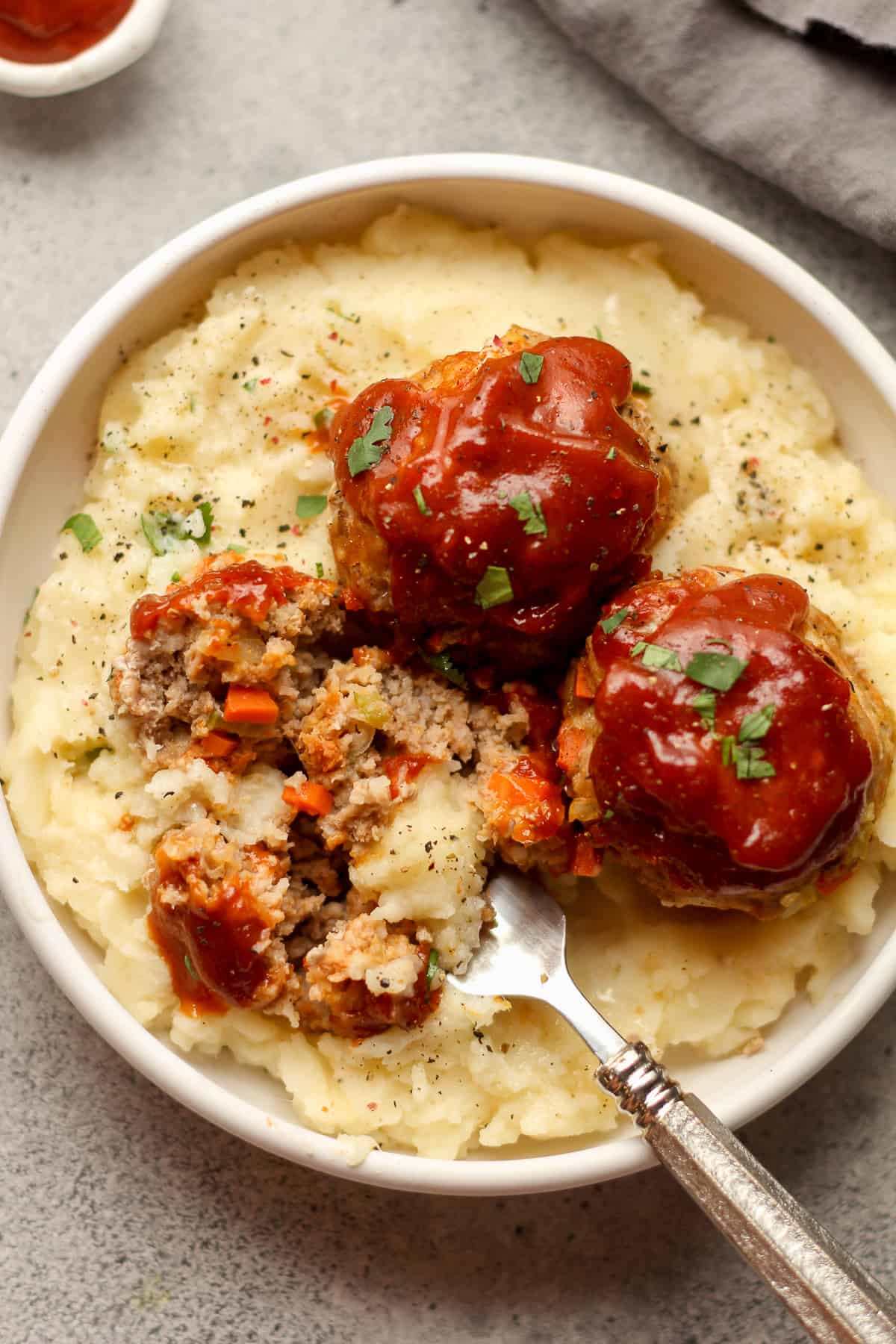 A closeup on a bowl of mashed potatoes with turkey meatloaf muffins on top, with sauce.