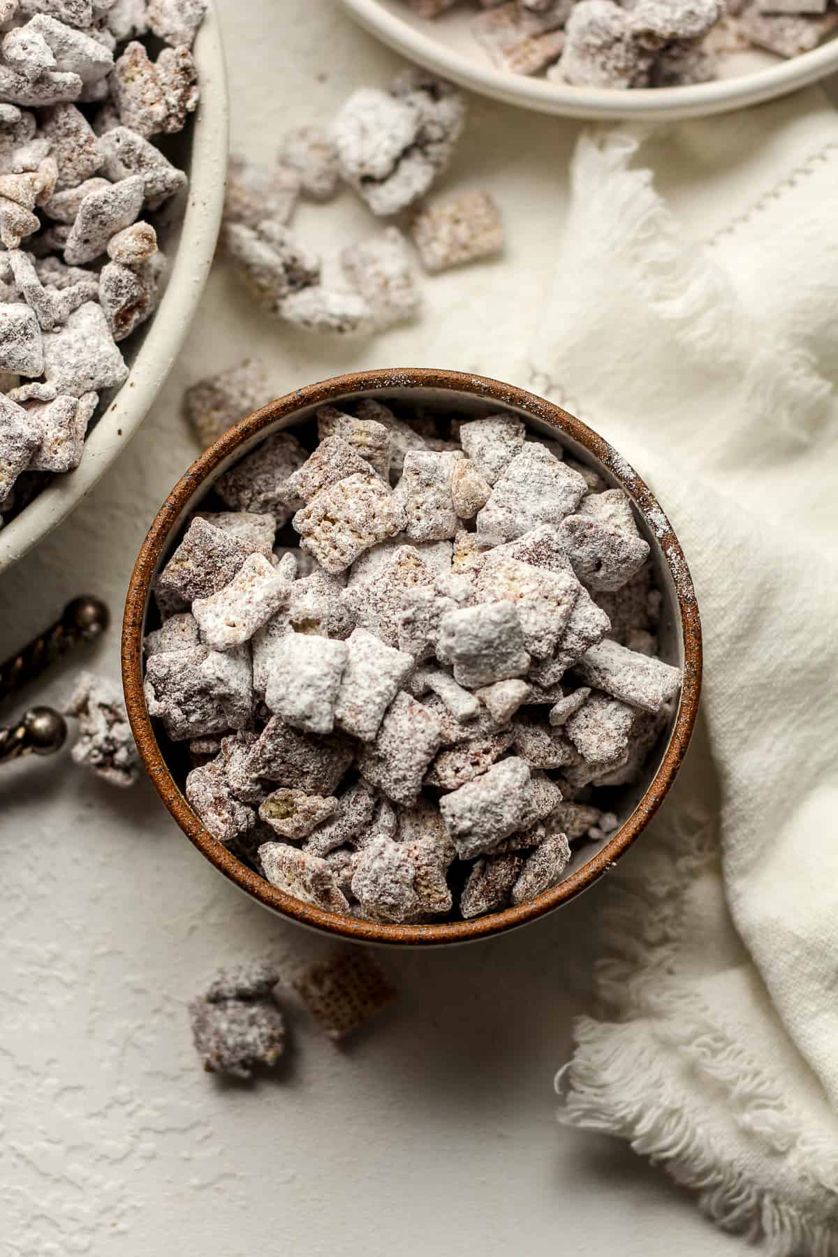 A small bowl of chocolate muddy buddies next to two other bowls and a white napkin.