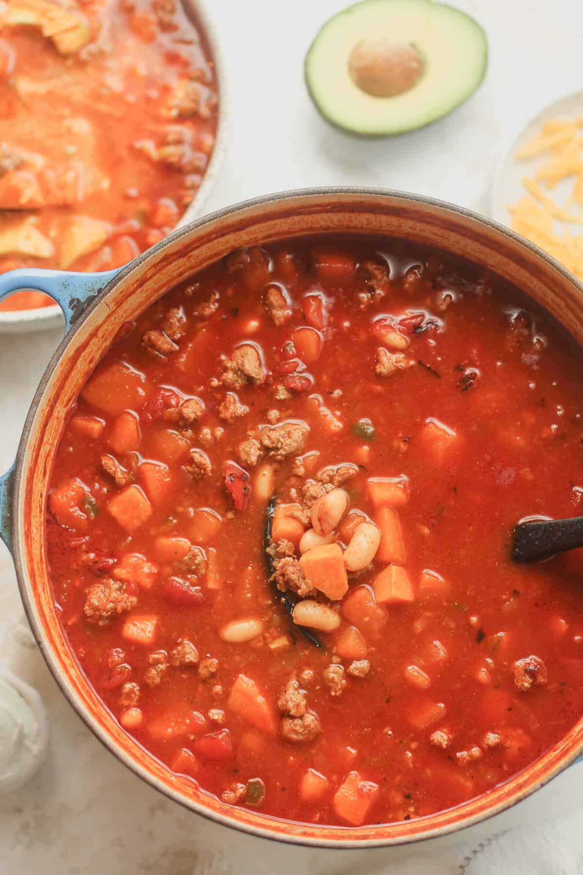 A large stock pot of sweet potato turkey chili next to a bowl of chili and toppings.