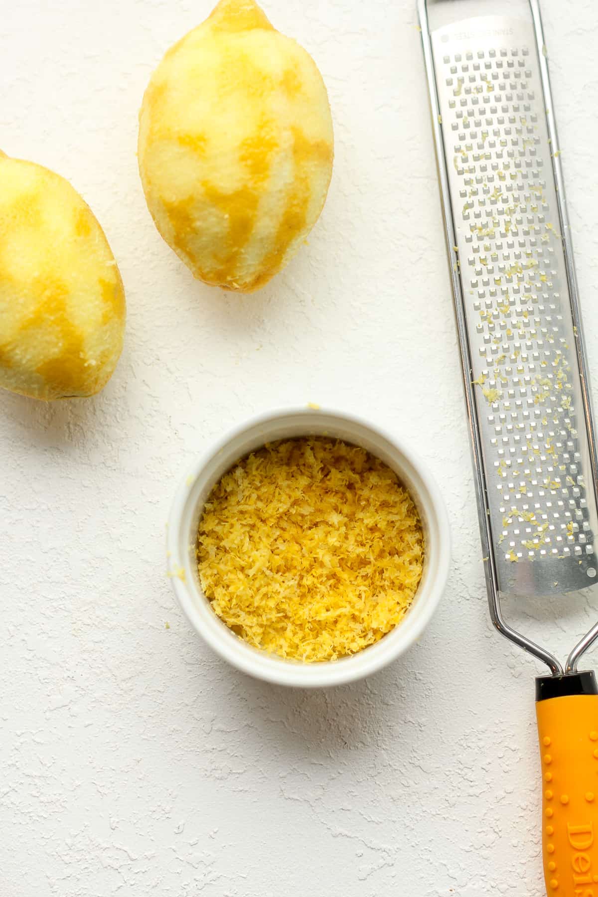 A bowl of lemon zest next to a lemon and a lemon zester.