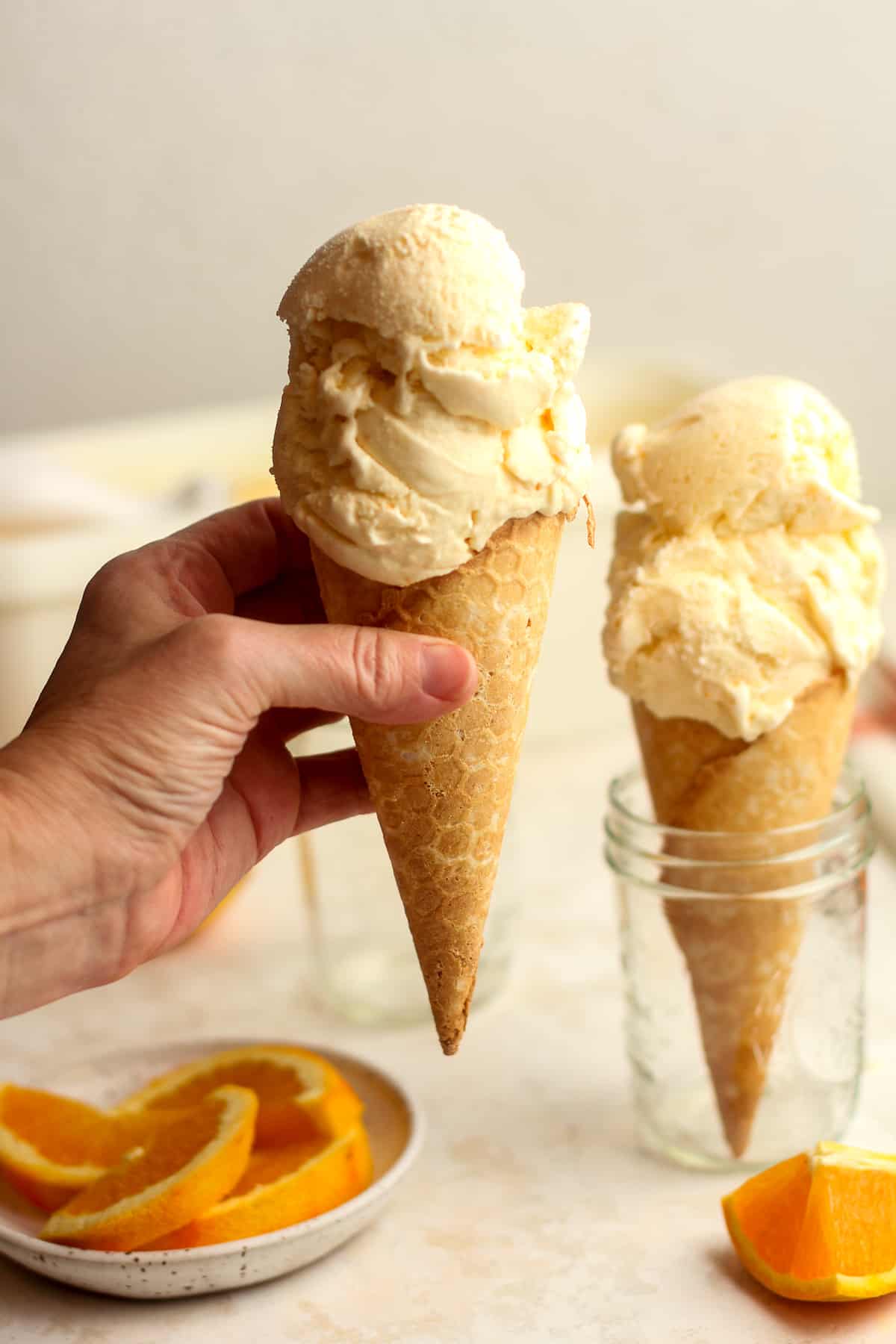 A hand holding an orange ice cream cone with another beside it.