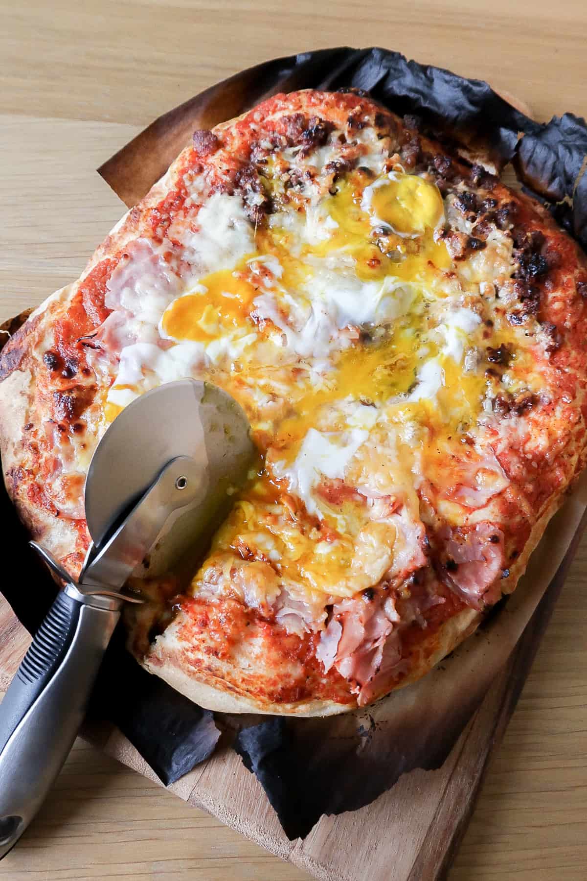A whole breakfast pizza on a board with a pizza cutter on top.