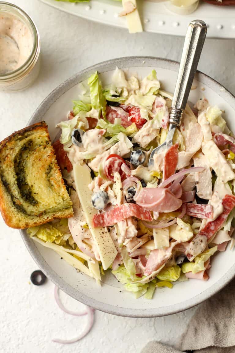 A shallow salad bowl with grinder salad mixed with salad dressing and topped with a slice of pesto bread.