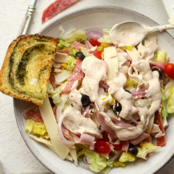 Closeup on a bowl of grinder salad with creamy dressing drizzled on top, and a piece of pesto bread.