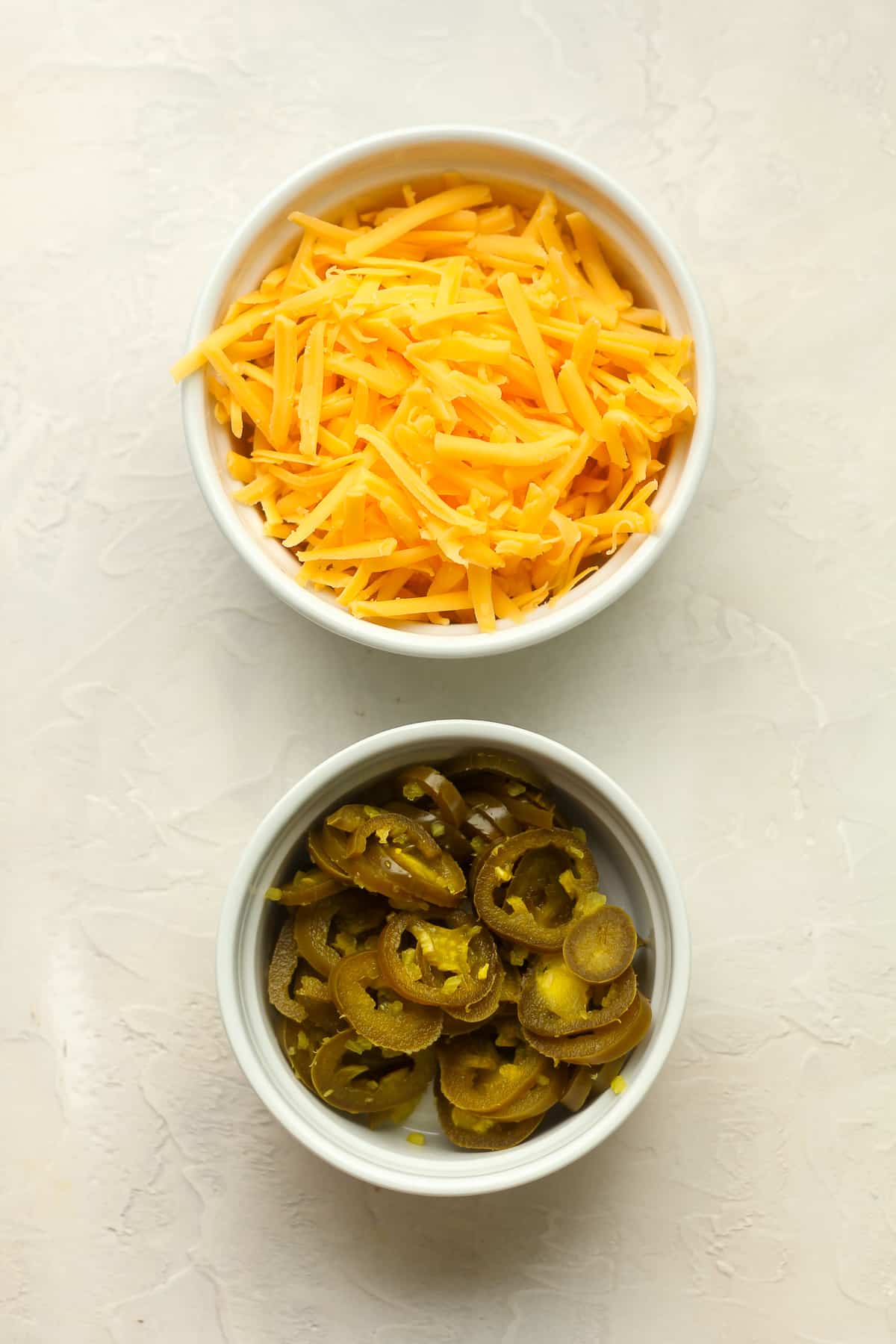 Two bowls - one with shredded cheddar cheese and the other with jarred jalapenos.
