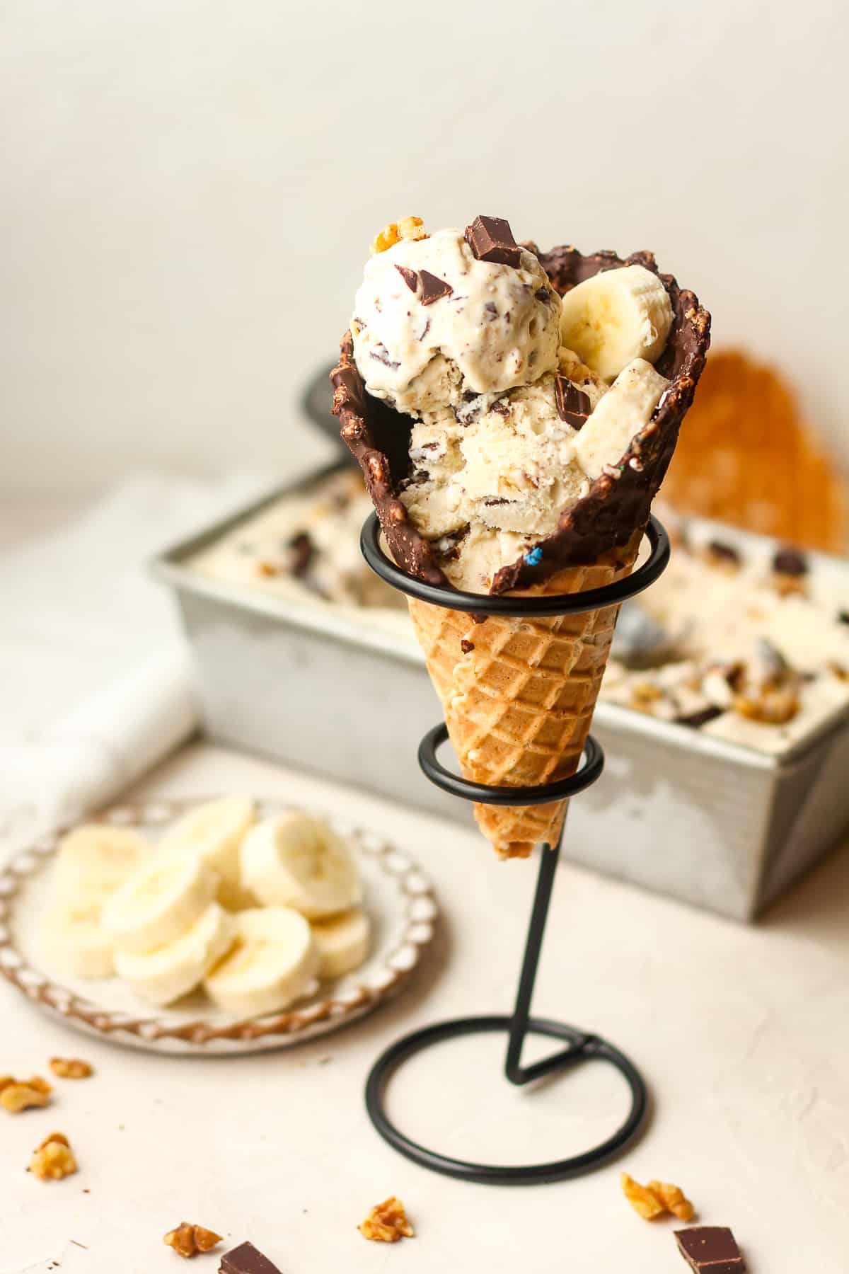 A chunky monkey ice cream cone with a rectangular pan of ice cream in the background.