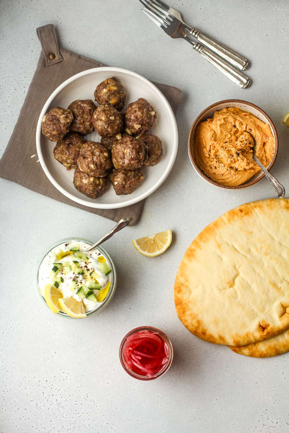 Overhead view of a bowl of meatballs, naan bread, and bowls of hummus and tzatziki sauce and a jar of pickled onions..