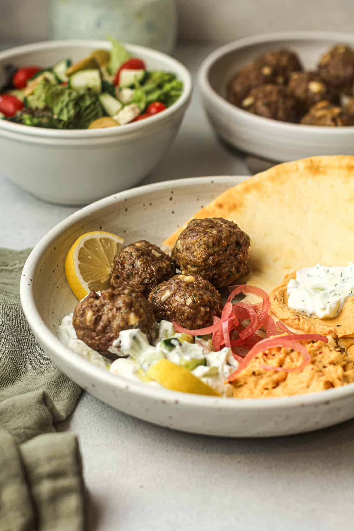 Side view of a bowl with naan, meatballs, tzatziki, and hummus - with a Greek salad in the background.