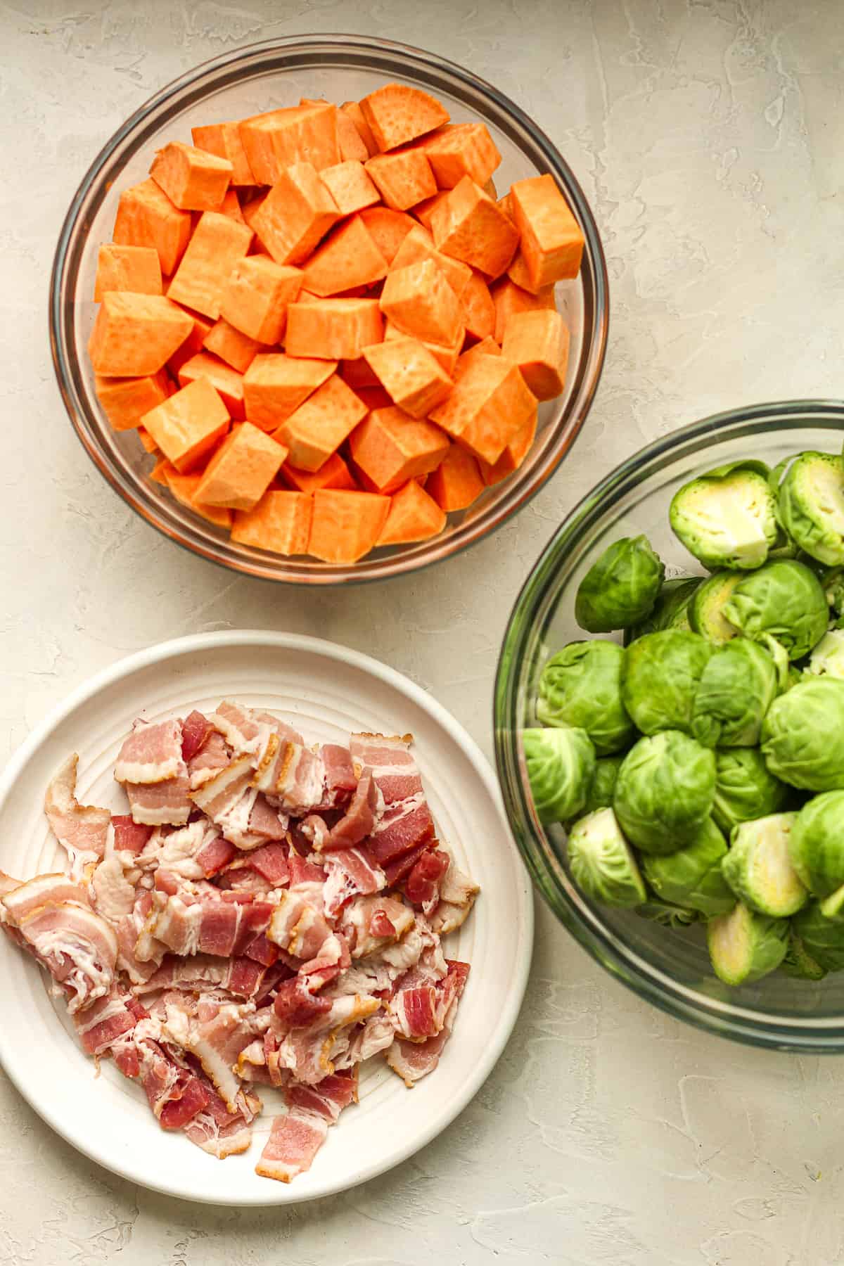 A bowl of chopped sweet potatoes, a bowl of halved Brussels sprouts, and a plate of chopped raw bacon.
