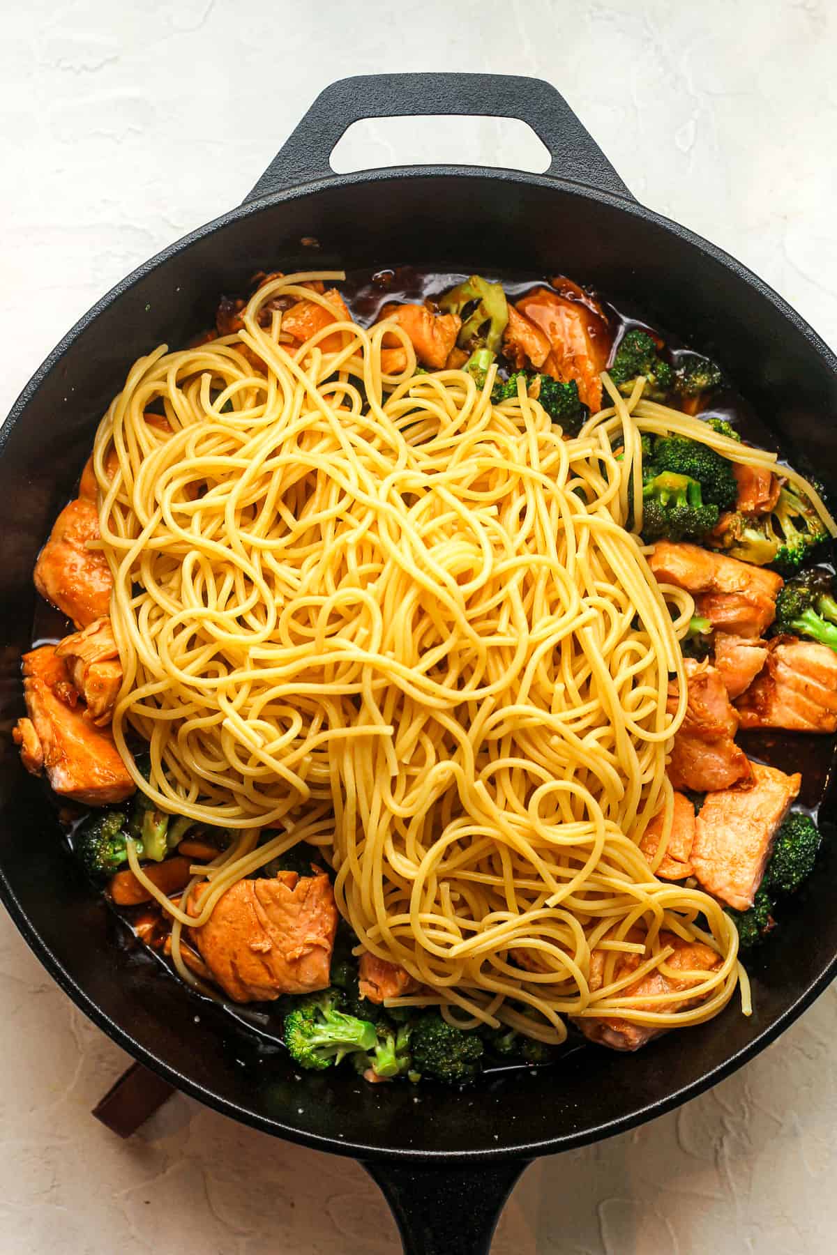 A skillet of teriyaki salmon and broccoli with noodles on top.