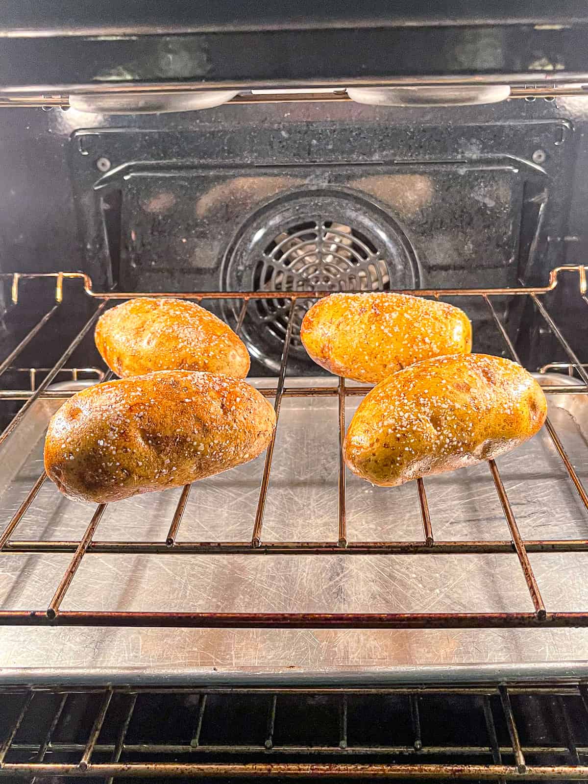 Four potatoes on the rack of an oven with a pan below it on another rack.