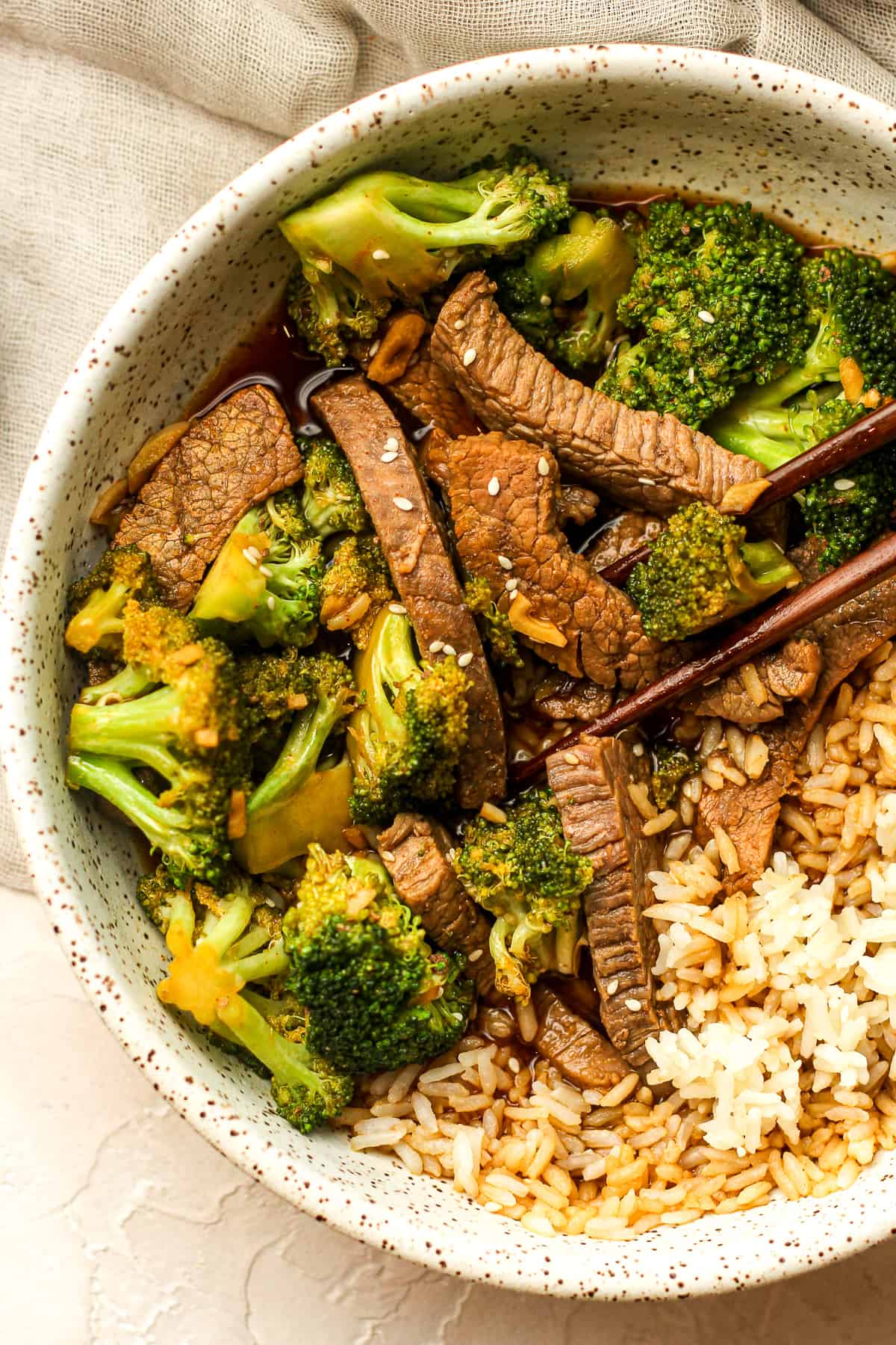 A bowl of beef and broccoli stir fry over rice with chop sticks.
