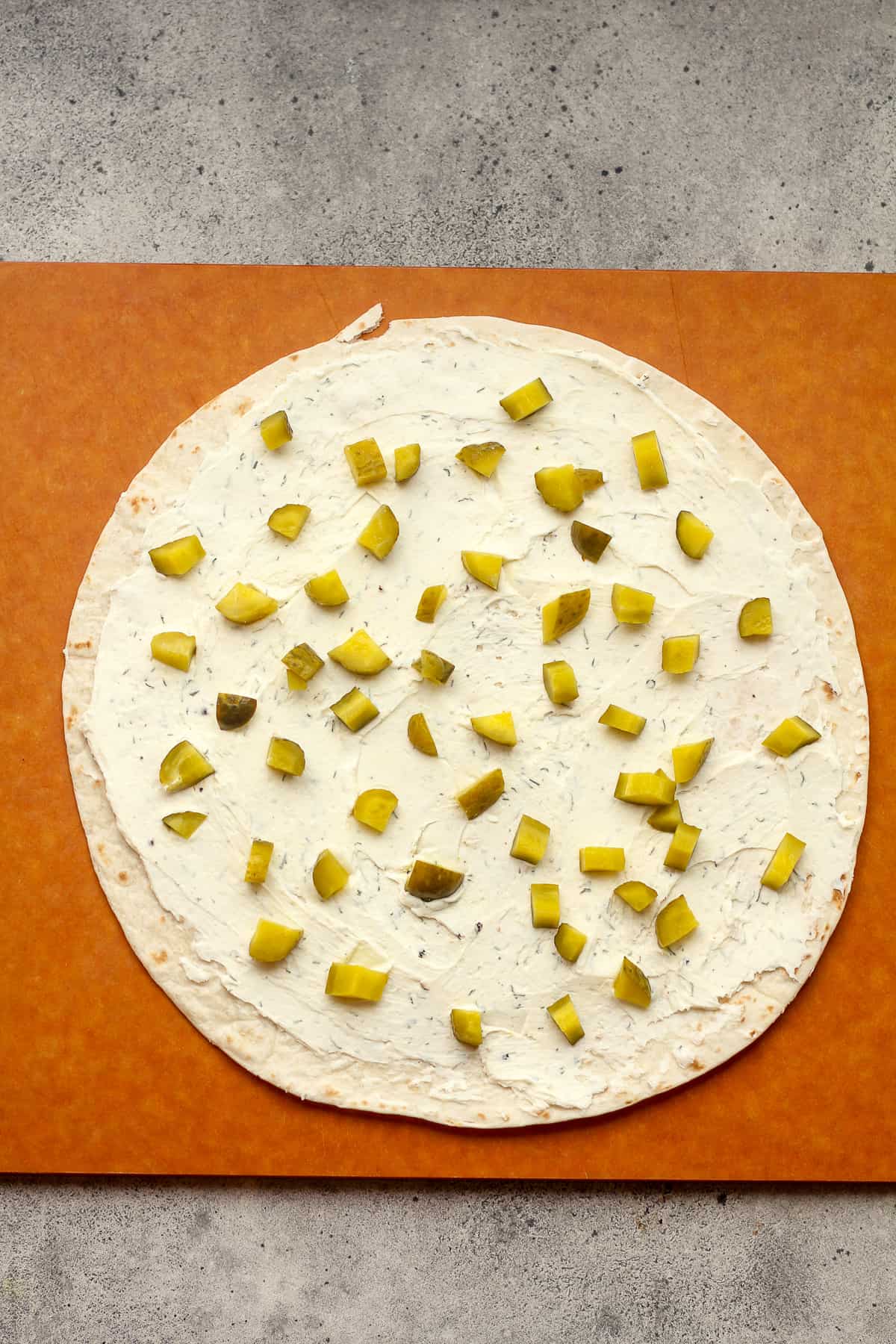 A large tortilla with cream cheese spread on top plus dill pickle pieces.