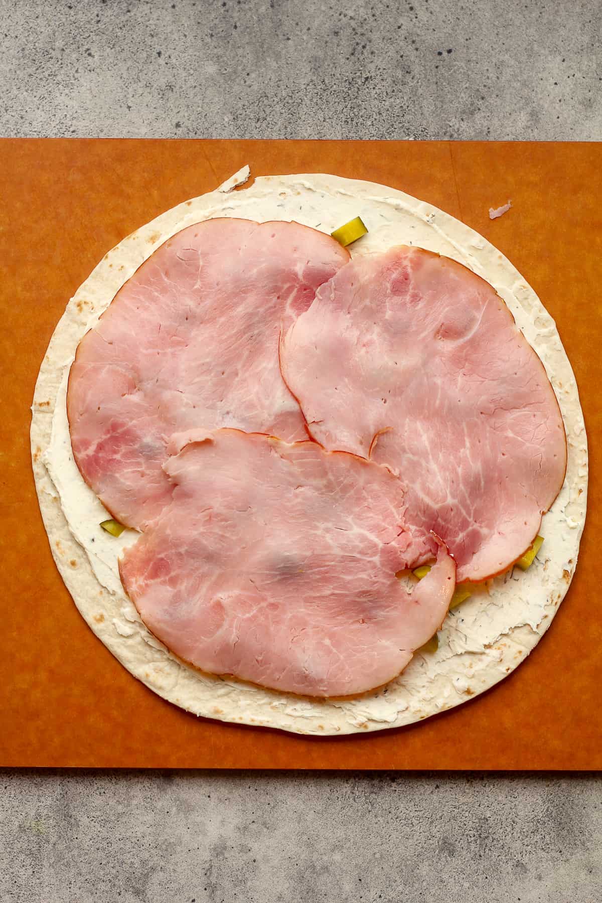 A cutting board with a large tortilla, cream cheese, pickles, plus ham on top.