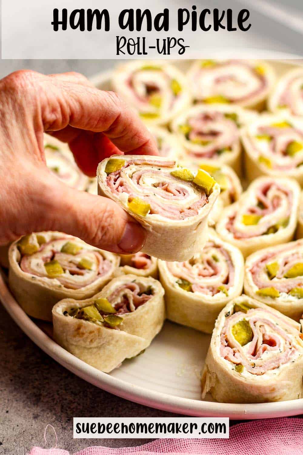 A hand holding a ham and pickle rollup over a plate of them.