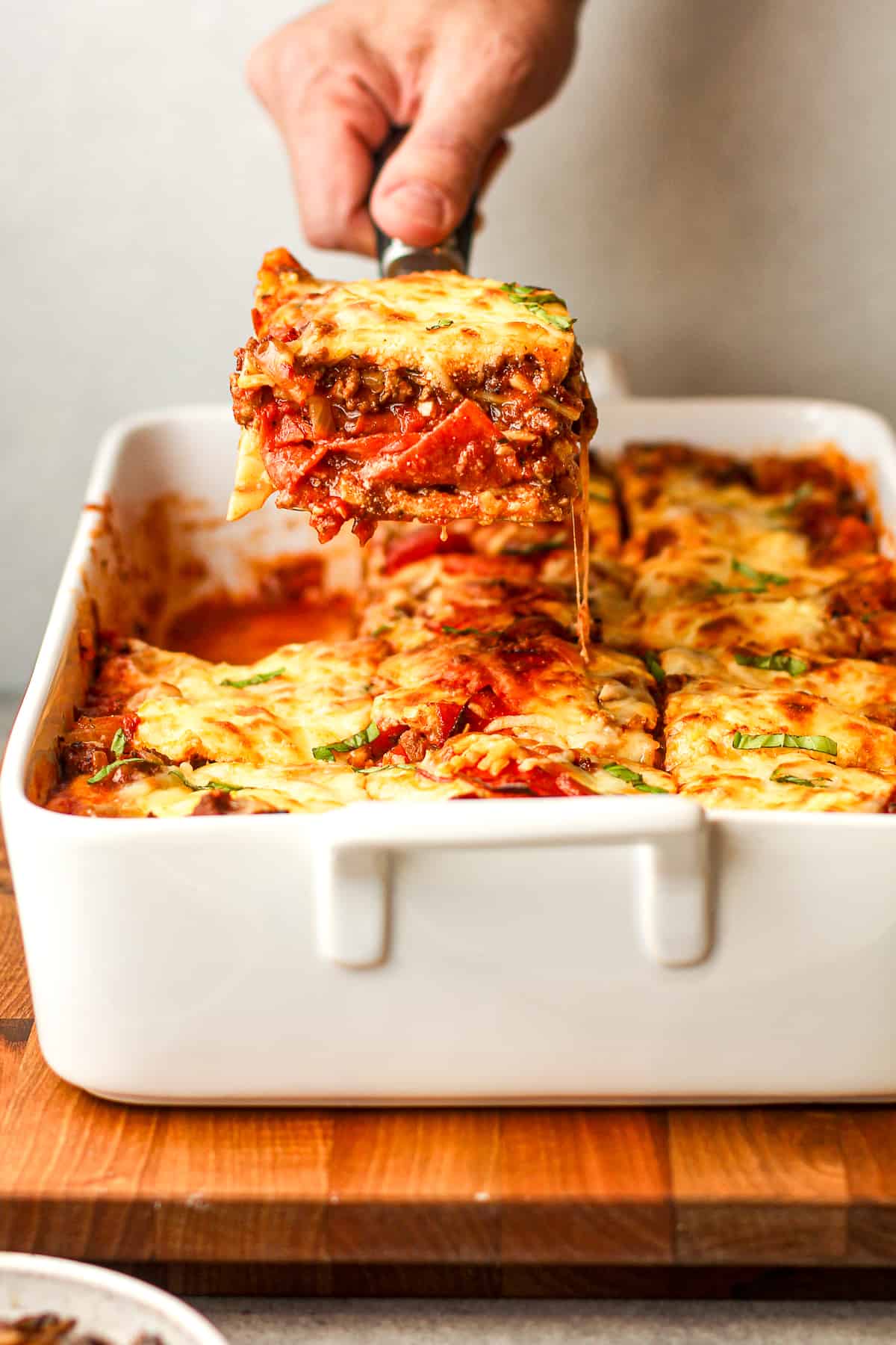 A hand lifting up a square of meat lovers lasagna over a casserole dish.