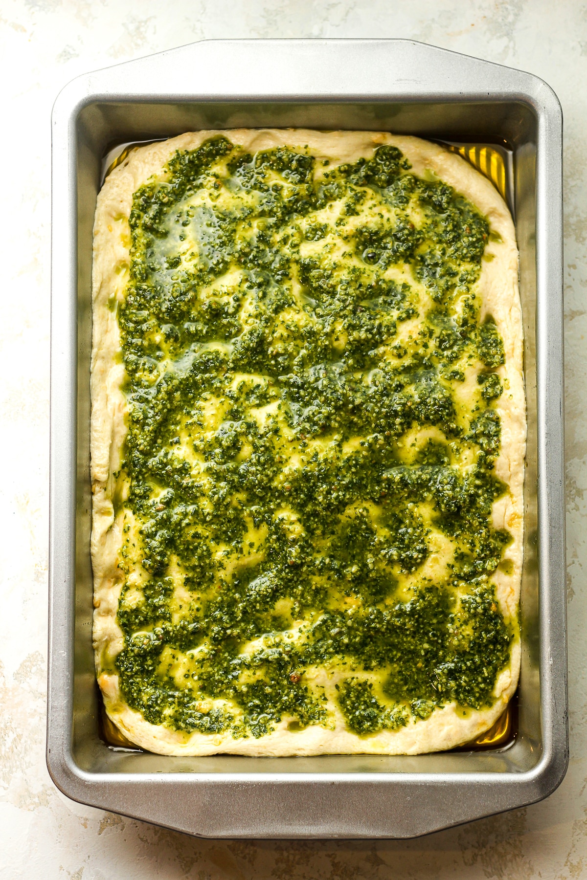 A pan of the focaccia dough with pesto sauce on top.