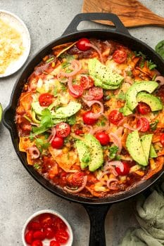 A skillet of beef enchiladas with sliced avocado and tomatoes on top.