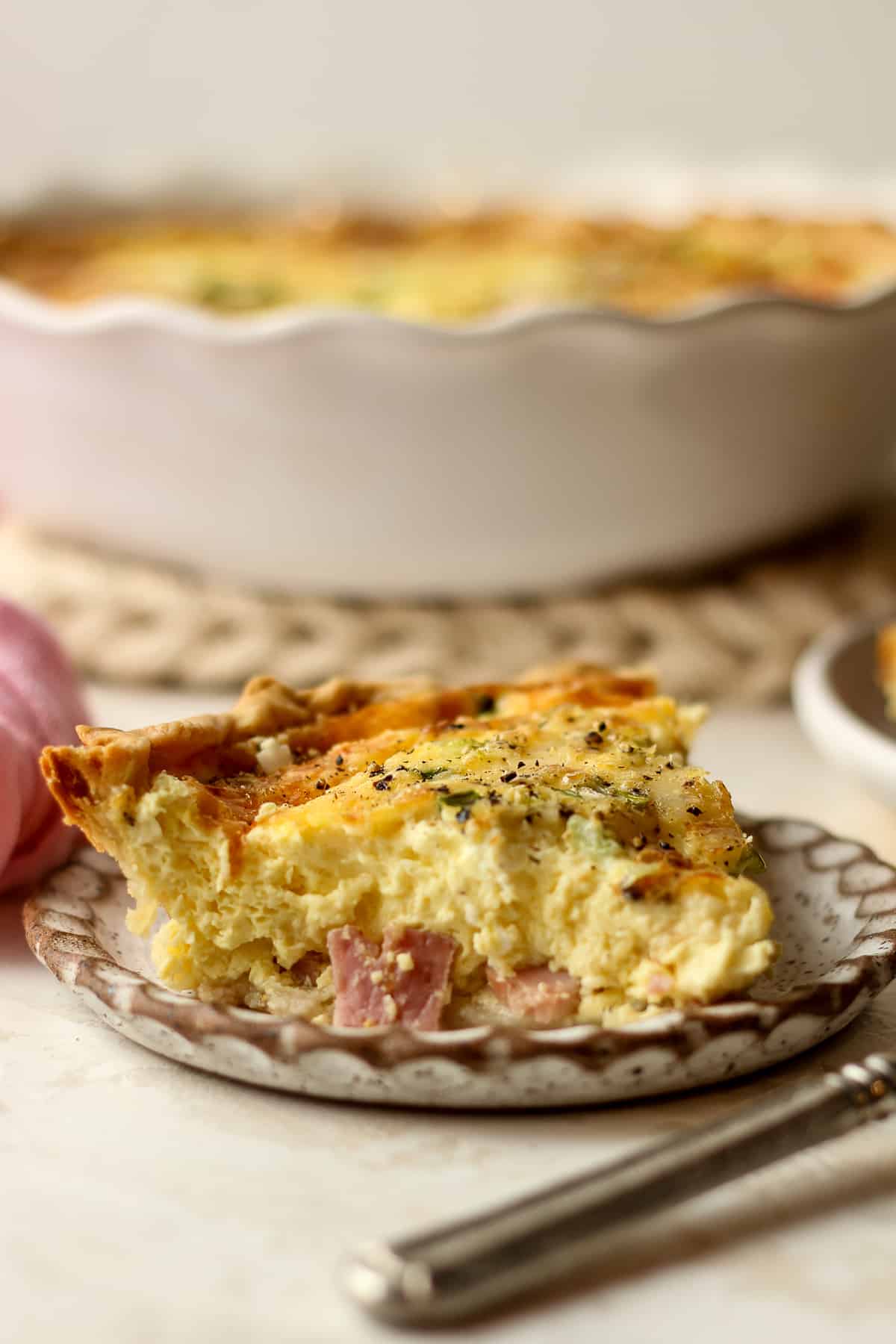 Side view of a slice of ham and gruyere quiche with the pie plate in the back.