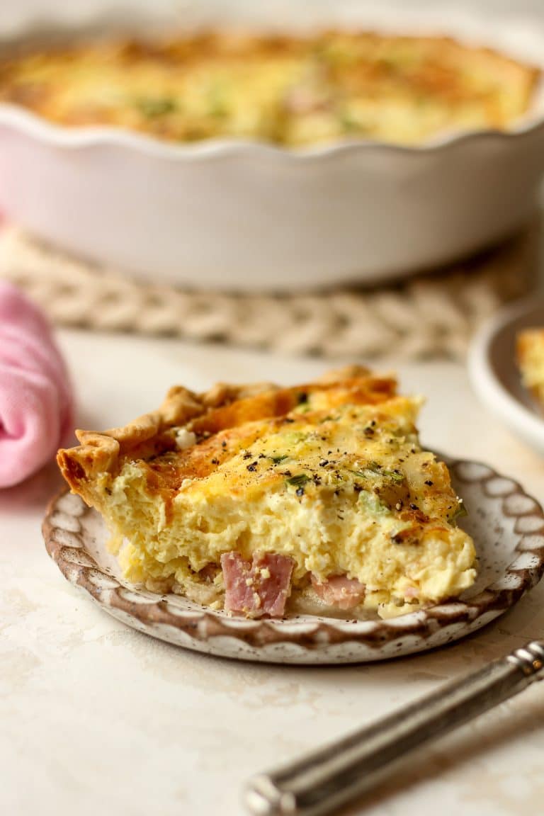 A slice of quiche with a pie plate of quiche in the background.