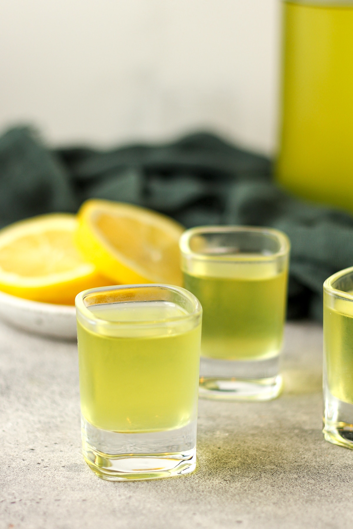 Side view of three shots of limoncello with lemon slices in the background.