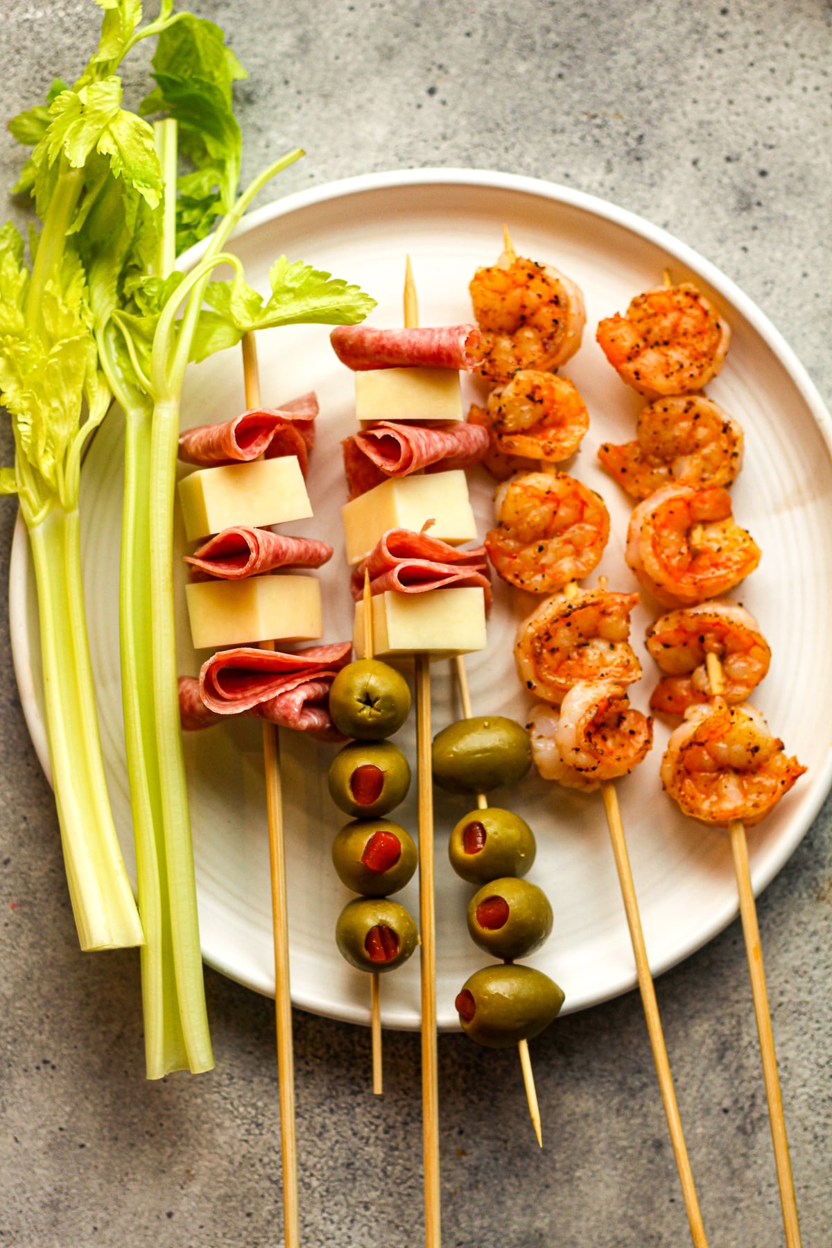 A plate of garnishes - celery, meat and salami skewer, olives, and shrimp.