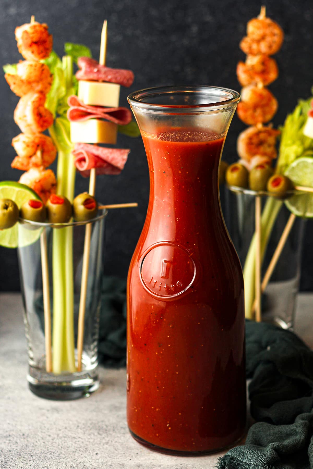 Side view of a large carafe of Bloody Mary drinks with glasses in the background.