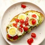 A white plate with a piece of burrata toast with avocado and egg.