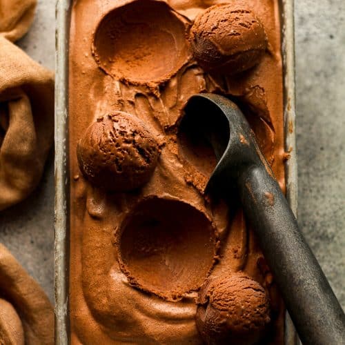 Closeup on a loaf pan of dark chocolate ice cream with several dips out and a scoop.