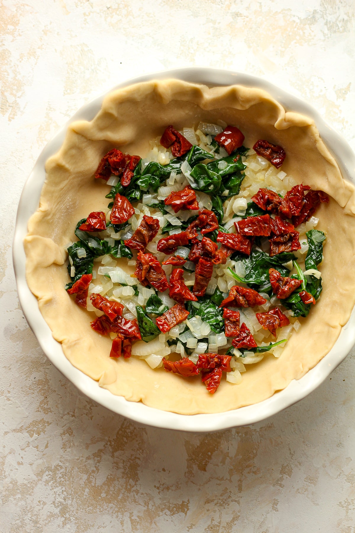 A pie plate with the crust and the veggies and sun-dried tomatoes.