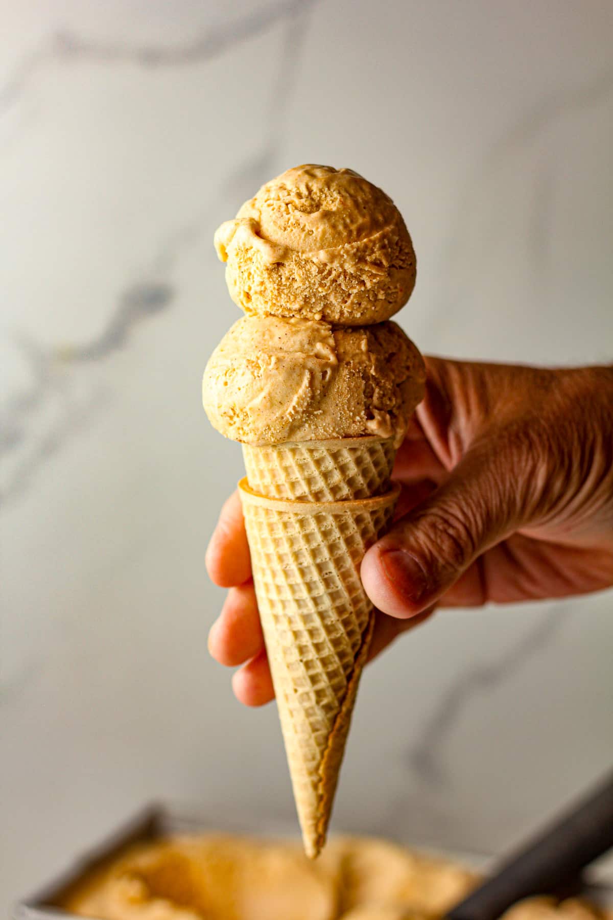 A hand holding a sugar cone with two scoops of pumpkin ice cream.