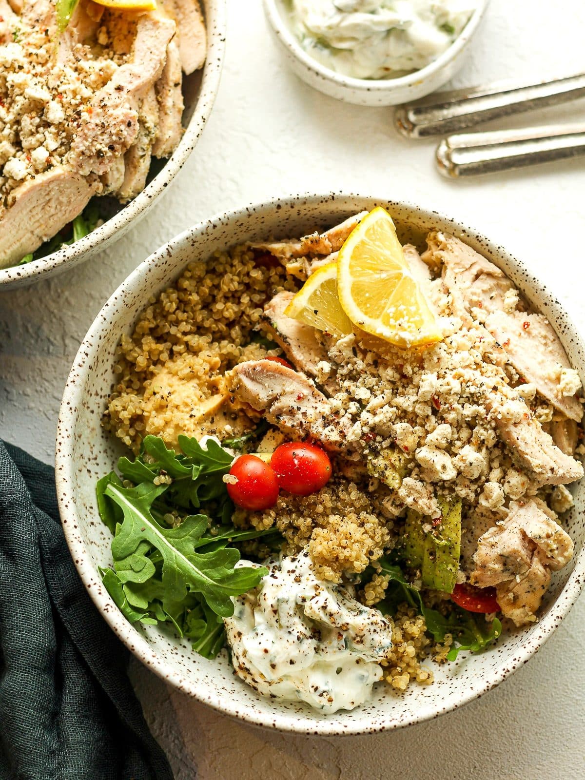 Two bowls of quinoa, Greek chicken, hummus, and tzatziki sauce.