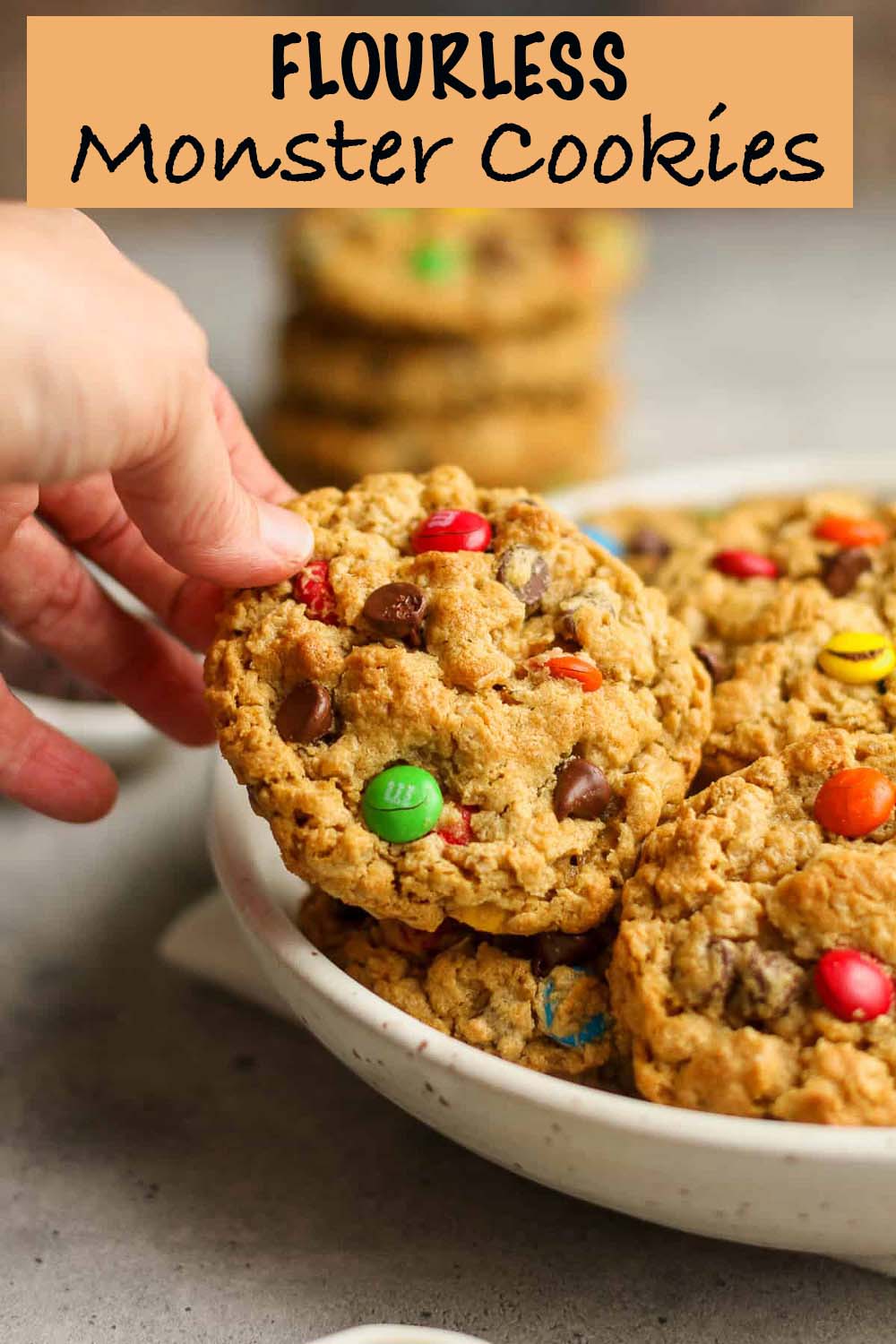 A hand reaching for a flourless monster cookie in a bowl.