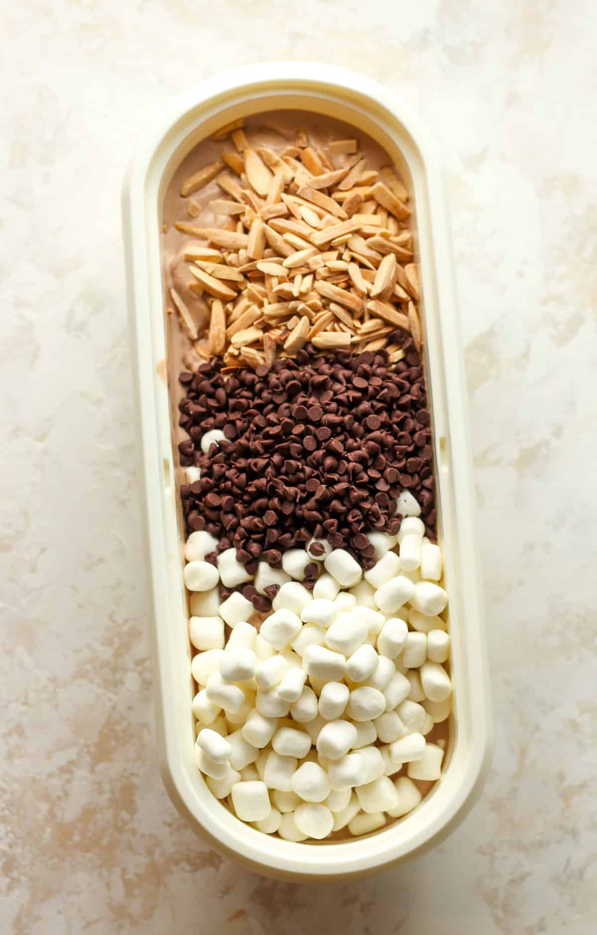 The ice cream container with chocolate ice cream topped with toasted almonds, mini chocolate chips, and mini marshmallows.