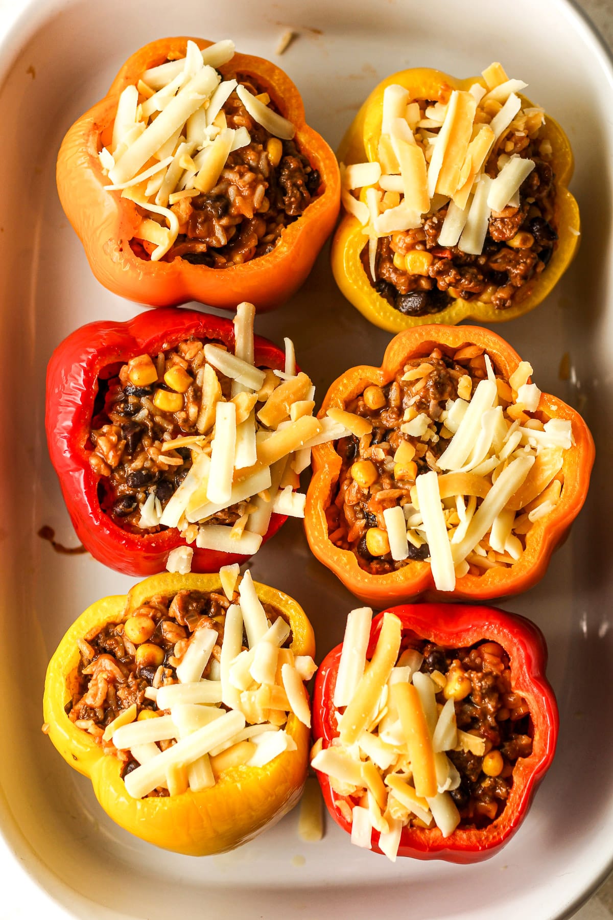 A casserole dish of six bell peppers stuffed with meat mixture and shredded cheese.