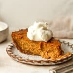 Side view of a slice of pumpkin pie in a graham cracker crust with a dollop of whipped cream.