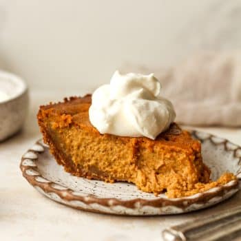 Side view of a slice of pumpkin pie in a graham cracker crust with a dollop of whipped cream.