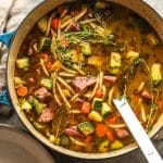 A pot of vegetable soup with ham and a soup ladle.