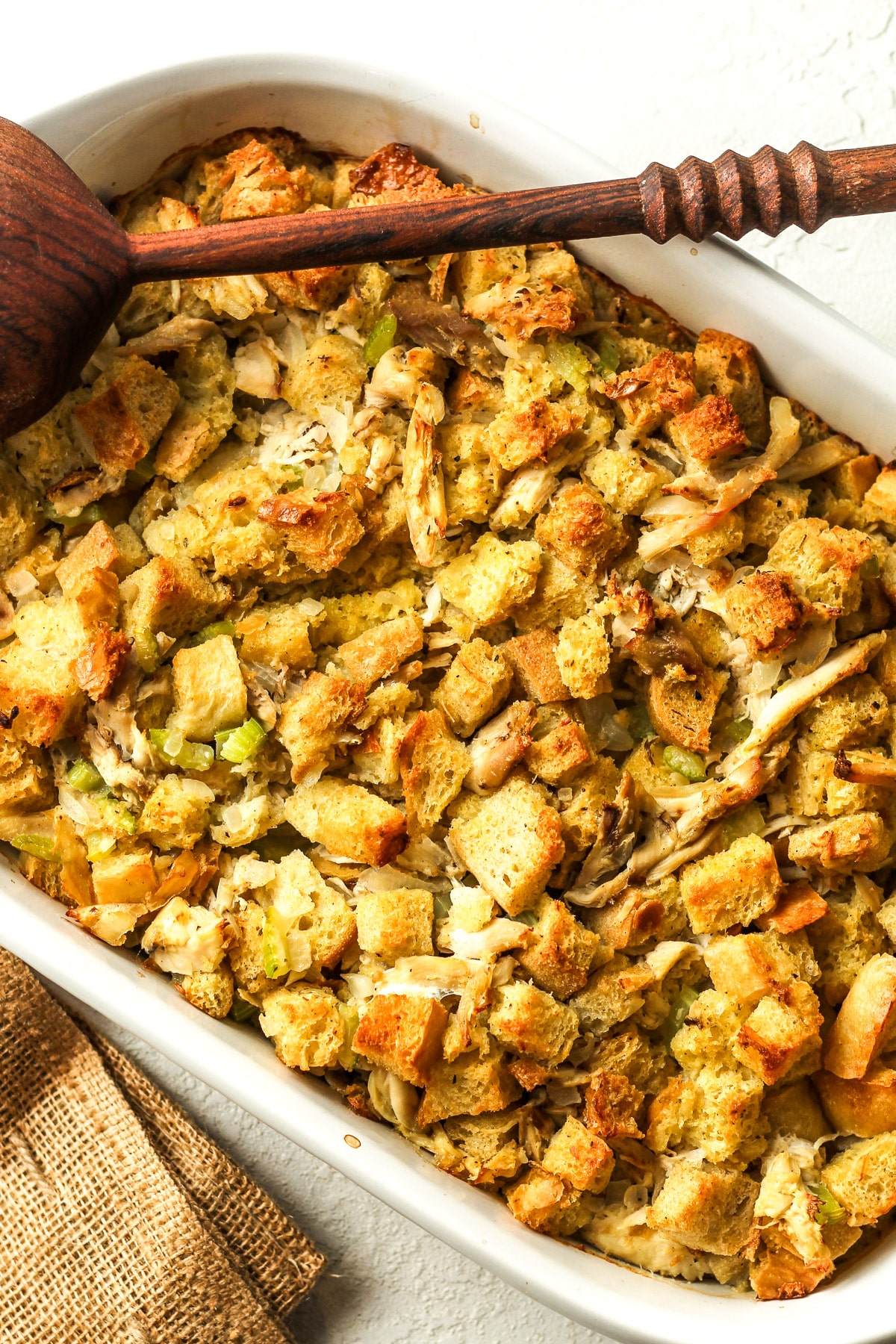 A tilted rectangular dish of Thanksgiving stuffing with a wooden spoon.