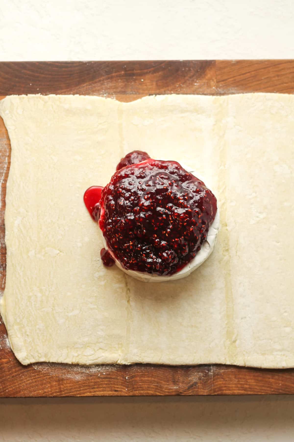 A sheet of puff pastry on a board topped with brie cheese and then raspberry jam.