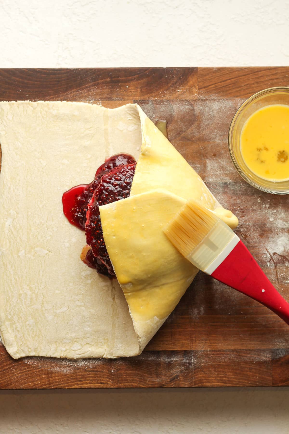 A board with the puff pastry being wrapped around the brie and jam showing how to add the egg wash.