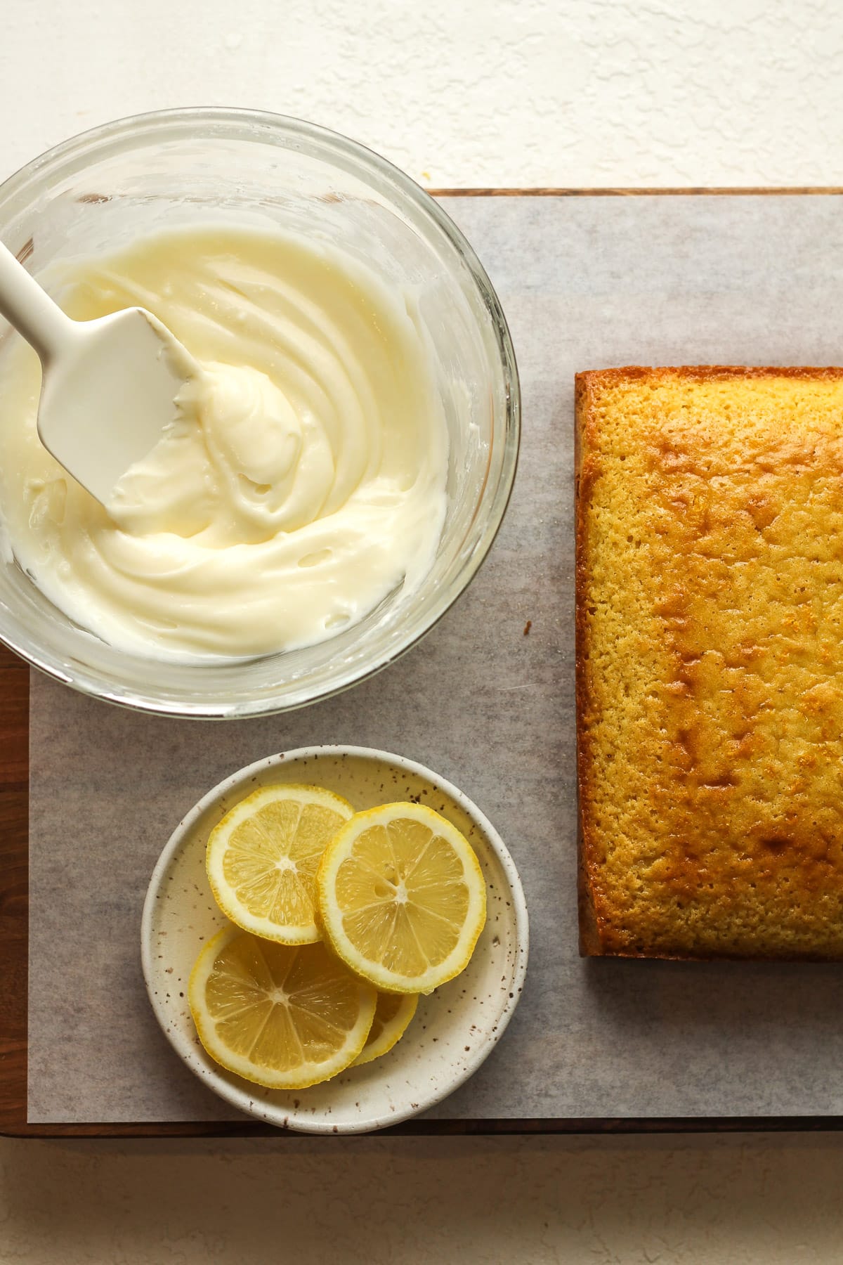 A board with a lemon cake, a bowl of the cream cheese icing, and a small plate of sliced lemon.