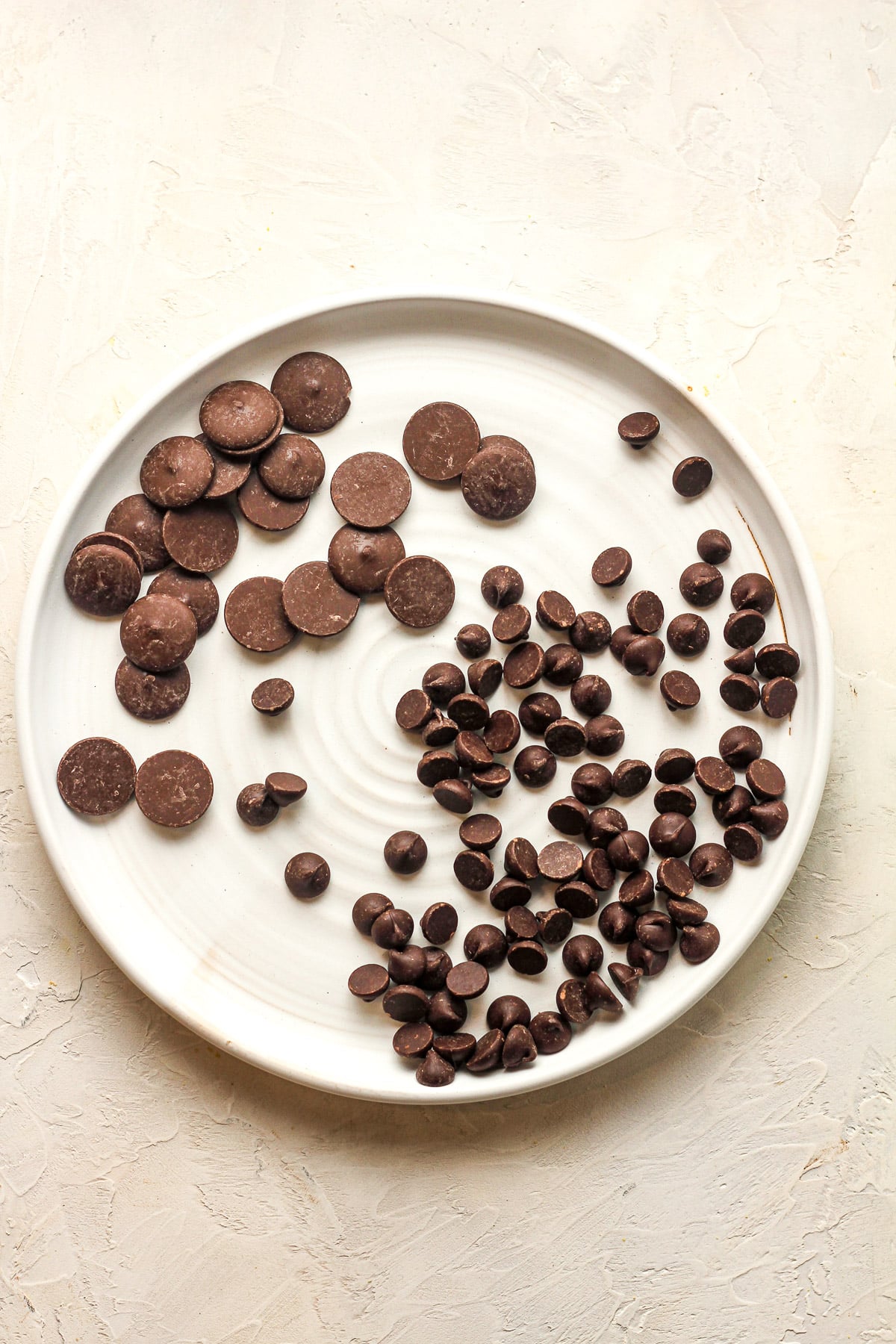 A white plate of dark chocolate chips and dark chocolate melting wafers.