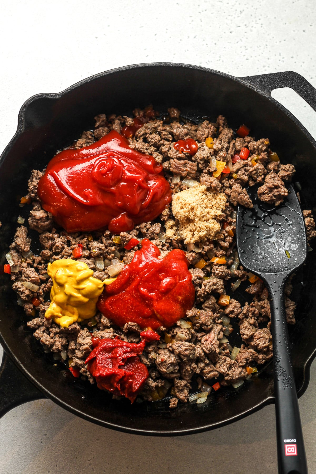 A skillet of the ground beef with ketchup, sriracha, mustard, tomato paste, and brown sugar on top.